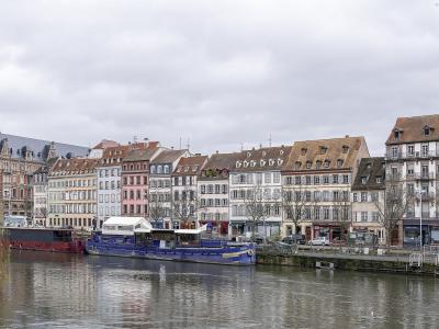 The image shows a row of multi-story buildings with varied facades (white, pink, beige) lining a riverbank, with a large blue barge docked in the foreground and a weeping willow on the left bank.