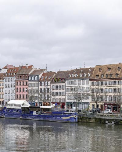 The image shows a row of multi-story buildings with varied facades (white, pink, beige) lining a riverbank, with a large blue barge docked in the foreground and a weeping willow on the left bank.