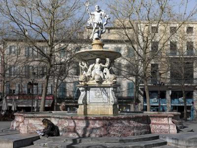 A large, ornate stone fountain with a statue of Neptune at the top, situated in a paved public square with bare trees and buildings in the background.