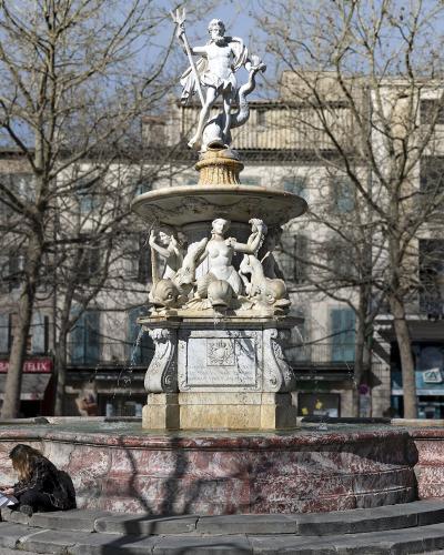 A large, ornate stone fountain with a statue of Neptune at the top, situated in a paved public square with bare trees and buildings in the background.