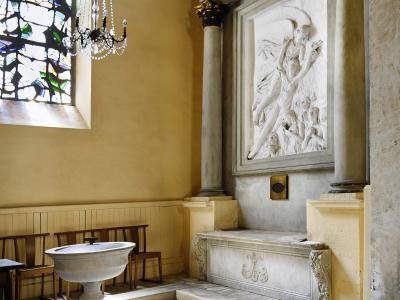 The image shows an interior view of a church featuring a stone baptismal font in the foreground, a stained-glass window to the left, and a large white marble relief sculpture set within a classical architectural niche on the right wall.