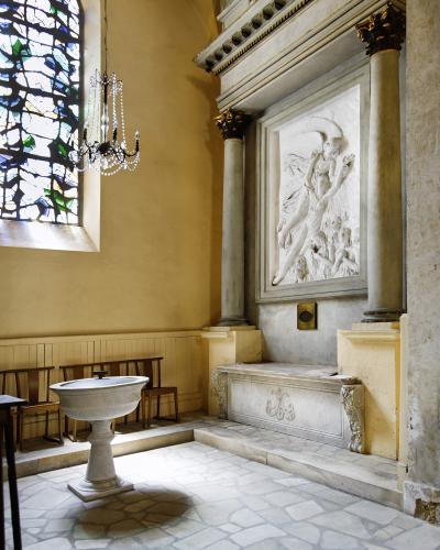 The image shows an interior view of a church featuring a stone baptismal font in the foreground, a stained-glass window to the left, and a large white marble relief sculpture set within a classical architectural niche on the right wall.