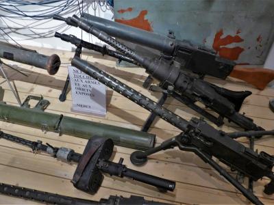 The image shows a museum display of various World War II-era firearms and weapons, including submachine guns and a rocket launcher, arranged on a wooden floor with a sign in French reading "NE PAS TOUCHER AUX ARMES ET AUX OBJETS EXPOSES MERCI" (Do