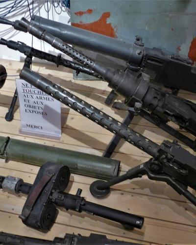 The image shows a museum display of various World War II-era firearms and weapons, including submachine guns and a rocket launcher, arranged on a wooden floor with a sign in French reading "NE PAS TOUCHER AUX ARMES ET AUX OBJETS EXPOSES MERCI" (Do