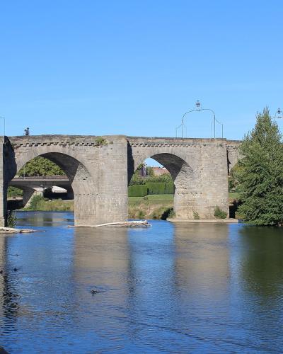 A multi-arched medieval stone bridge spanning a wide river under a clear blue sky, with pedestrians walking on top and modern street lamps mounted along the parapet; a newer concrete bridge is visible in the background to the left.