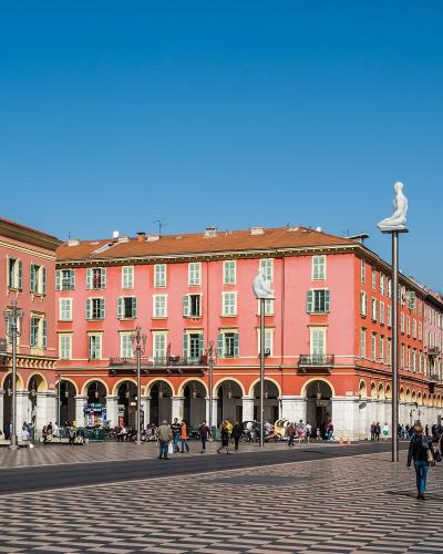 A wide-angle daytime view of a large European plaza with distinctive black-and-white checkered paving, flanked by colorful buildings with arched ground-floor colonnades and topped by white statues of seated figures on tall poles.