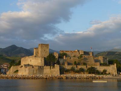 A stone fortress complex situated on a rocky promontory overlooking a bay, with mountains in the background and a flag flying from one of its towers.