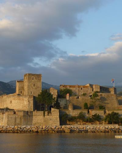 A stone fortress complex situated on a rocky promontory overlooking a bay, with mountains in the background and a flag flying from one of its towers.