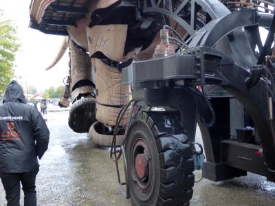 A low-angle outdoor shot of the rear and side of the Grand Éléphant mechanical sculpture in Nantes, showing its massive wooden body, large black tires, metal framework, and a security guard wearing a jacket labeled "SÉCURITÉ PRIVÉE LYNX ASSISTANCE