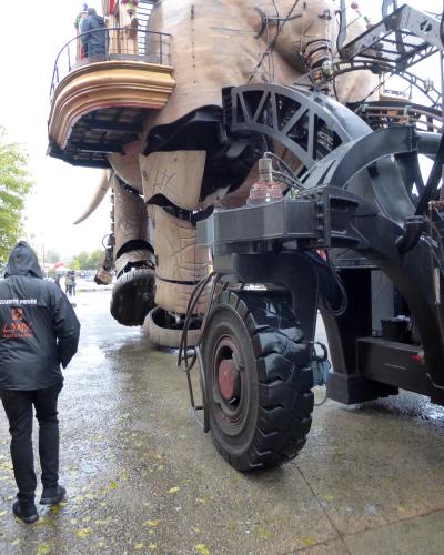 A low-angle outdoor shot of the rear and side of the Grand Éléphant mechanical sculpture in Nantes, showing its massive wooden body, large black tires, metal framework, and a security guard wearing a jacket labeled "SÉCURITÉ PRIVÉE LYNX ASSISTANCE
