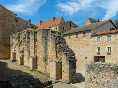 The image shows the ruins of ancient stone walls and foundations, likely part of a historical complex, with adjacent buildings featuring red-tiled roofs and white-framed windows under a blue sky.