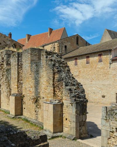 The image shows the ruins of ancient stone walls and foundations, likely part of a historical complex, with adjacent buildings featuring red-tiled roofs and white-framed windows under a blue sky.