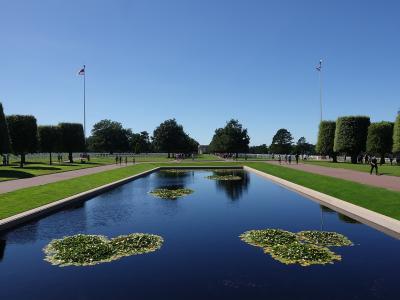 A long rectangular reflecting pool with lily pads, flanked by manicured lawns, pathways, and rows of white headstones under a clear blue sky, with two American flags on tall poles.