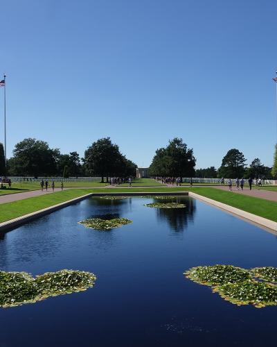 A long rectangular reflecting pool with lily pads, flanked by manicured lawns, pathways, and rows of white headstones under a clear blue sky, with two American flags on tall poles.