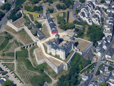 An aerial photograph showing a large medieval castle complex with multiple towers, fortified walls, and an adjacent courtyard, situated on a hill overlooking a town with vineyards and roads in the foreground.