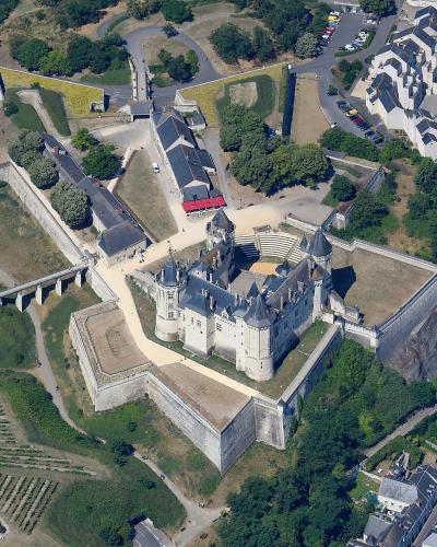 An aerial photograph showing a large medieval castle complex with multiple towers, fortified walls, and an adjacent courtyard, situated on a hill overlooking a town with vineyards and roads in the foreground.