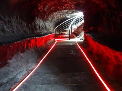 A dark, tunnel-like interior with rough rock walls, illuminated by red LED strip lights along the floor and leading toward a metal staircase at the far end.