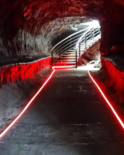 A dark, tunnel-like interior with rough rock walls, illuminated by red LED strip lights along the floor and leading toward a metal staircase at the far end.