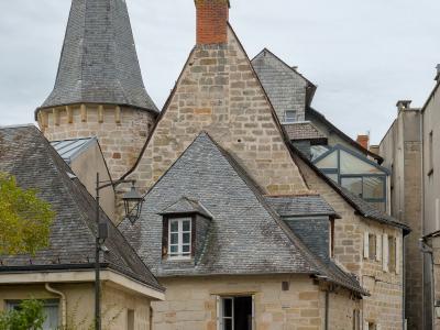 A street scene showing historic stone buildings with slate roofs, including a prominent conical turret, and parked cars along the road.