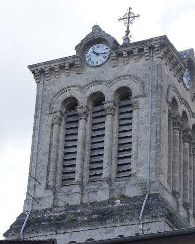 A stone bell tower with arched louvers, a clock face, and a cross on top, set against a cloudy sky.