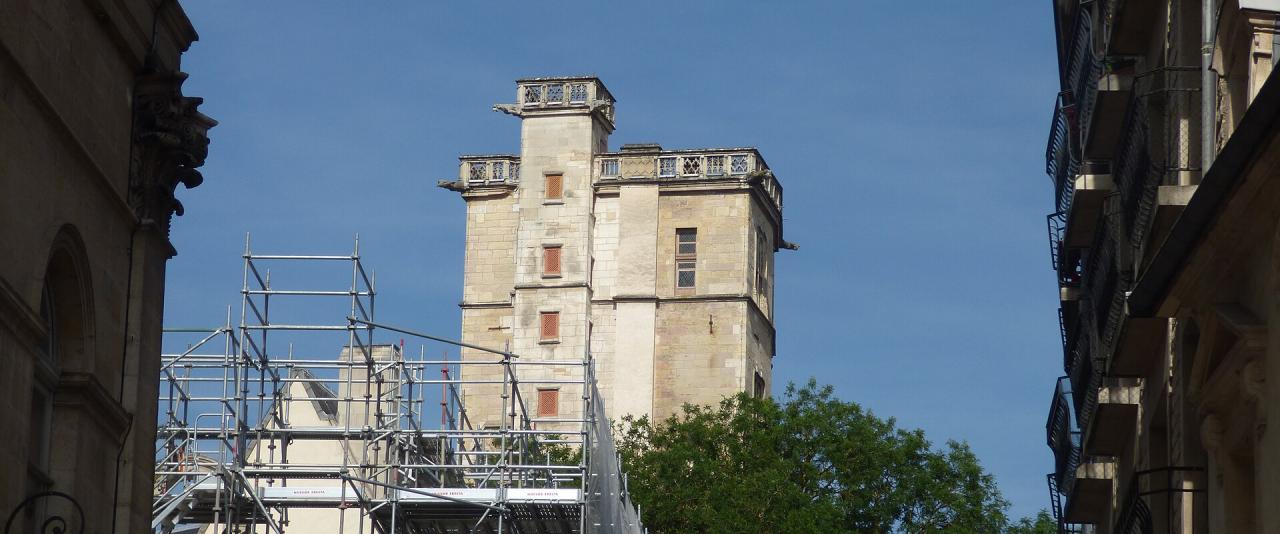 A stone tower with crenellated battlements and small windows, viewed from a narrow street between buildings, with scaffolding in the foreground and a clear blue sky above.