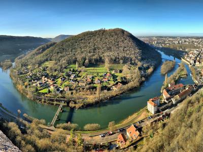 A panoramic view from an elevated stone rampart looking down at a large, U-shaped river meander enclosing a forested hill with scattered houses and a small village, with a city visible in the background under a clear blue sky.