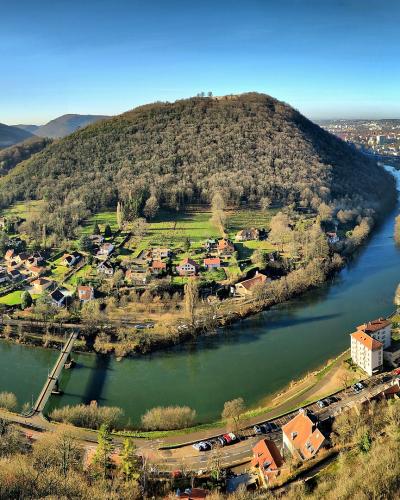 A panoramic view from an elevated stone rampart looking down at a large, U-shaped river meander enclosing a forested hill with scattered houses and a small village, with a city visible in the background under a clear blue sky.