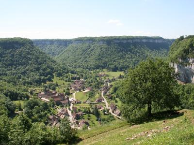 A panoramic view of a small village nestled in a steep, forested valley with dramatic limestone cliffs, featuring traditional stone houses and a prominent church or abbey complex.