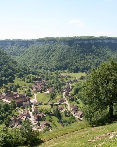 A panoramic view of a small village nestled in a steep, forested valley with dramatic limestone cliffs, featuring traditional stone houses and a prominent church or abbey complex.