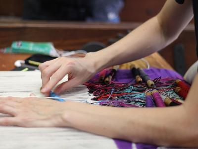 A close-up view of a person’s hands working on a tapestry loom, with colorful threads and wooden shuttles visible on a work surface.