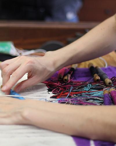 A close-up view of a person’s hands working on a tapestry loom, with colorful threads and wooden shuttles visible on a work surface.