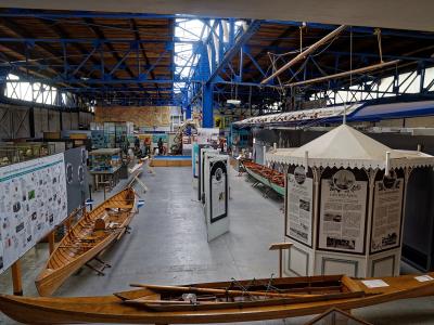 Interior of a large industrial warehouse converted into a museum, featuring wooden rowing shells, a model boat mast, and exhibition panels with French text such as "LES REGATES" and "LES CROISIERS".