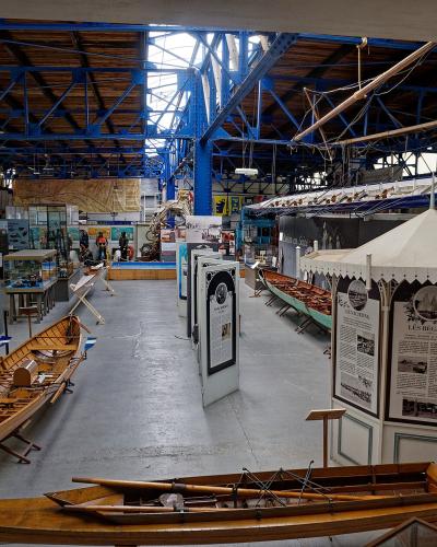 Interior of a large industrial warehouse converted into a museum, featuring wooden rowing shells, a model boat mast, and exhibition panels with French text such as "LES REGATES" and "LES CROISIERS".
