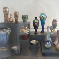 1. A museum display case containing a collection of colorful, ornate enamel vases and decorative objects in Art Nouveau style, arranged on tiered black pedestals behind glass.