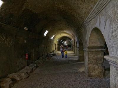 A long, vaulted underground stone corridor with arched openings on the right, small high windows on the left, scattered ancient stone fragments along the floor, and visitors walking through; a red fire extinguisher is mounted on the left wall.