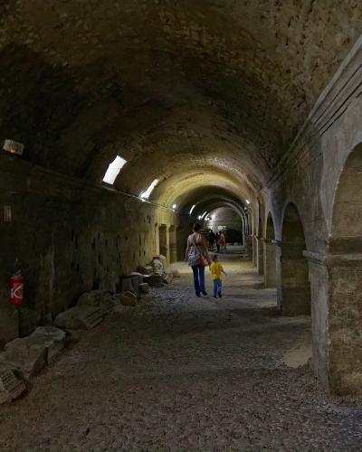 A long, vaulted underground stone corridor with arched openings on the right, small high windows on the left, scattered ancient stone fragments along the floor, and visitors walking through; a red fire extinguisher is mounted on the left wall.