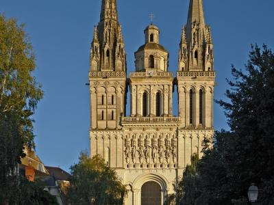 A low-angle photograph of the stone facade of a large Gothic cathedral featuring two tall spires, a central clock tower, and a row of statues above the main entrance, with temporary construction fencing and a white tent structure at its base.
