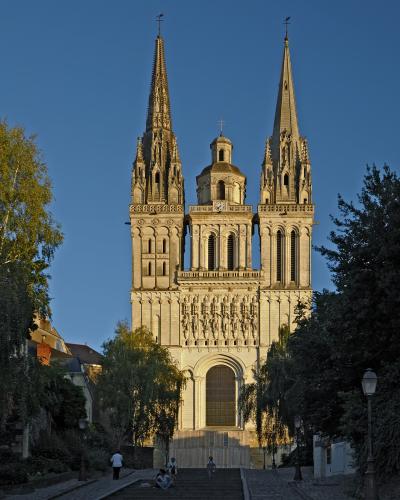 A low-angle photograph of the stone facade of a large Gothic cathedral featuring two tall spires, a central clock tower, and a row of statues above the main entrance, with temporary construction fencing and a white tent structure at its base.