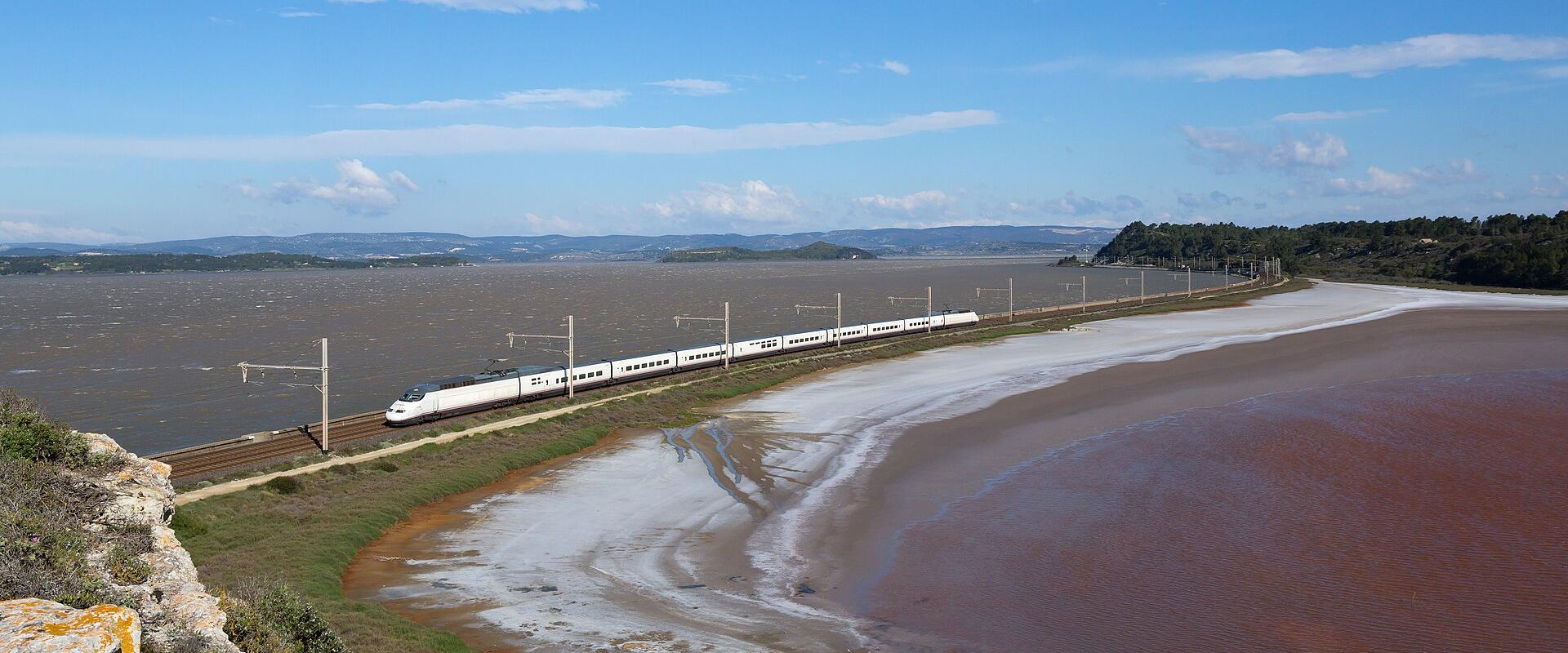 A white high-speed train travels along a coastal railway track beside a large body of reddish-brown water under a blue sky with scattered clouds.