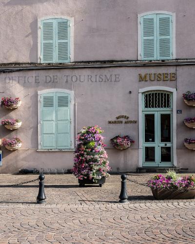 A pink building facade with light blue shutters, displaying signage for "OFFICE DE TOURISME" and "MUSEE MARIUS AUDIN," adorned with flower baskets and situated on a cobblestone street.