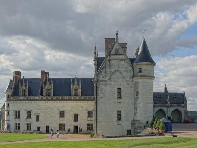 The image shows a large, historic French château with distinctive Renaissance architecture, including a prominent conical-roofed tower, ornate stone chimneys, and a long wing with decorative gables, set against a cloudy sky with visitors on the gr