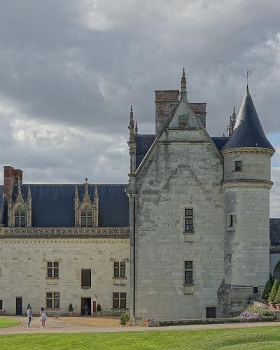 The image shows a large, historic French château with distinctive Renaissance architecture, including a prominent conical-roofed tower, ornate stone chimneys, and a long wing with decorative gables, set against a cloudy sky with visitors on the gr