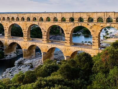 A panoramic photograph of a massive three-tiered Roman stone aqueduct bridge spanning a river valley, with people visible walking along the top tier and dense green vegetation on the banks.