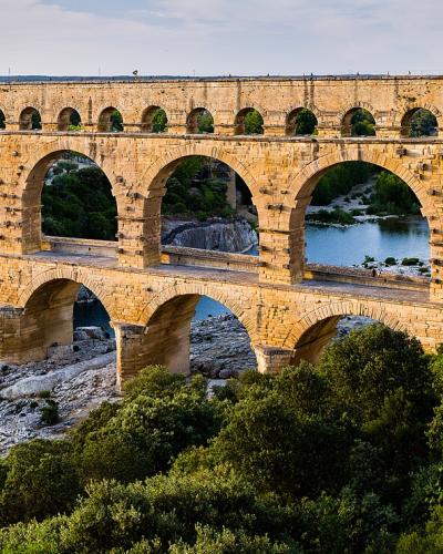 A panoramic photograph of a massive three-tiered Roman stone aqueduct bridge spanning a river valley, with people visible walking along the top tier and dense green vegetation on the banks.