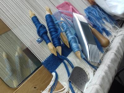 A close-up photograph of traditional low-warp tapestry weaving tools—including wooden shuttles (flûtes) wound with blue yarn, a metal comb (peigne), a beater (grattoir), and a punch tool (poinçon)—arranged on the warp threads of a wooden loom with