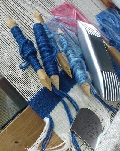 A close-up photograph of traditional low-warp tapestry weaving tools—including wooden shuttles (flûtes) wound with blue yarn, a metal comb (peigne), a beater (grattoir), and a punch tool (poinçon)—arranged on the warp threads of a wooden loom with