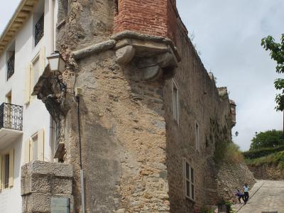 The image shows a section of old stone fortifications with a brick tower, adjacent to a white building and a cobblestone street, with a signpost in the foreground.