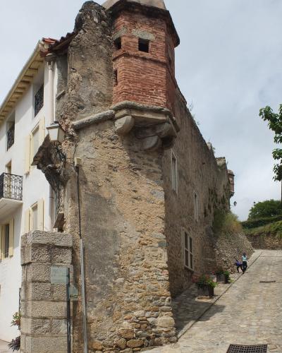 The image shows a section of old stone fortifications with a brick tower, adjacent to a white building and a cobblestone street, with a signpost in the foreground.