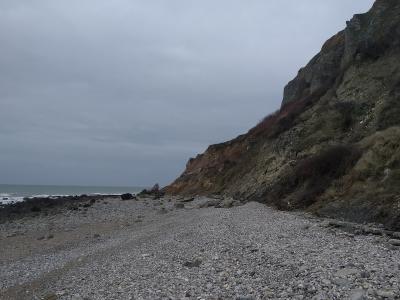 A pebble beach at the base of a steep, eroded cliff face under an overcast sky, with the sea visible to the left.