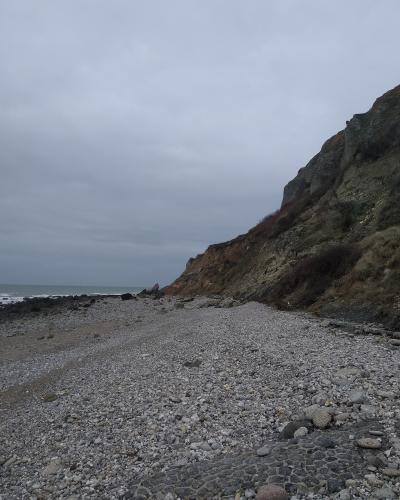 A pebble beach at the base of a steep, eroded cliff face under an overcast sky, with the sea visible to the left.